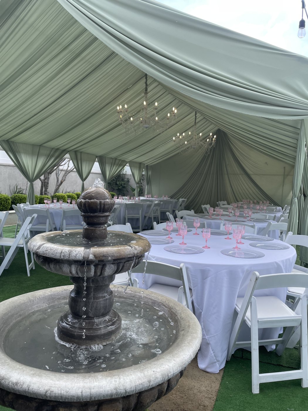 Elegant garden reception under a sage-green draped tent with chandeliers, a stone fountain, white round tables and pink glassware